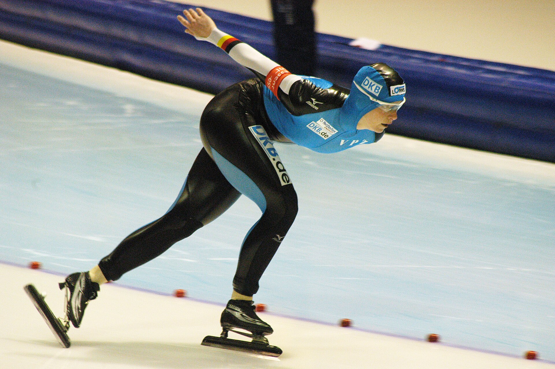 An action photograph of a speed skater in a blue and black racing suit mid-stride on an ice rink. The skater is leaned forward in a low, aerodynamic crouch, showing the specialized clap skate in action: the heel of the left boot is lifted significantly off the long metal blade, which remains flat against the ice thanks to a hinge at the toe. The background shows the blurred blue padding of the rink's perimeter.