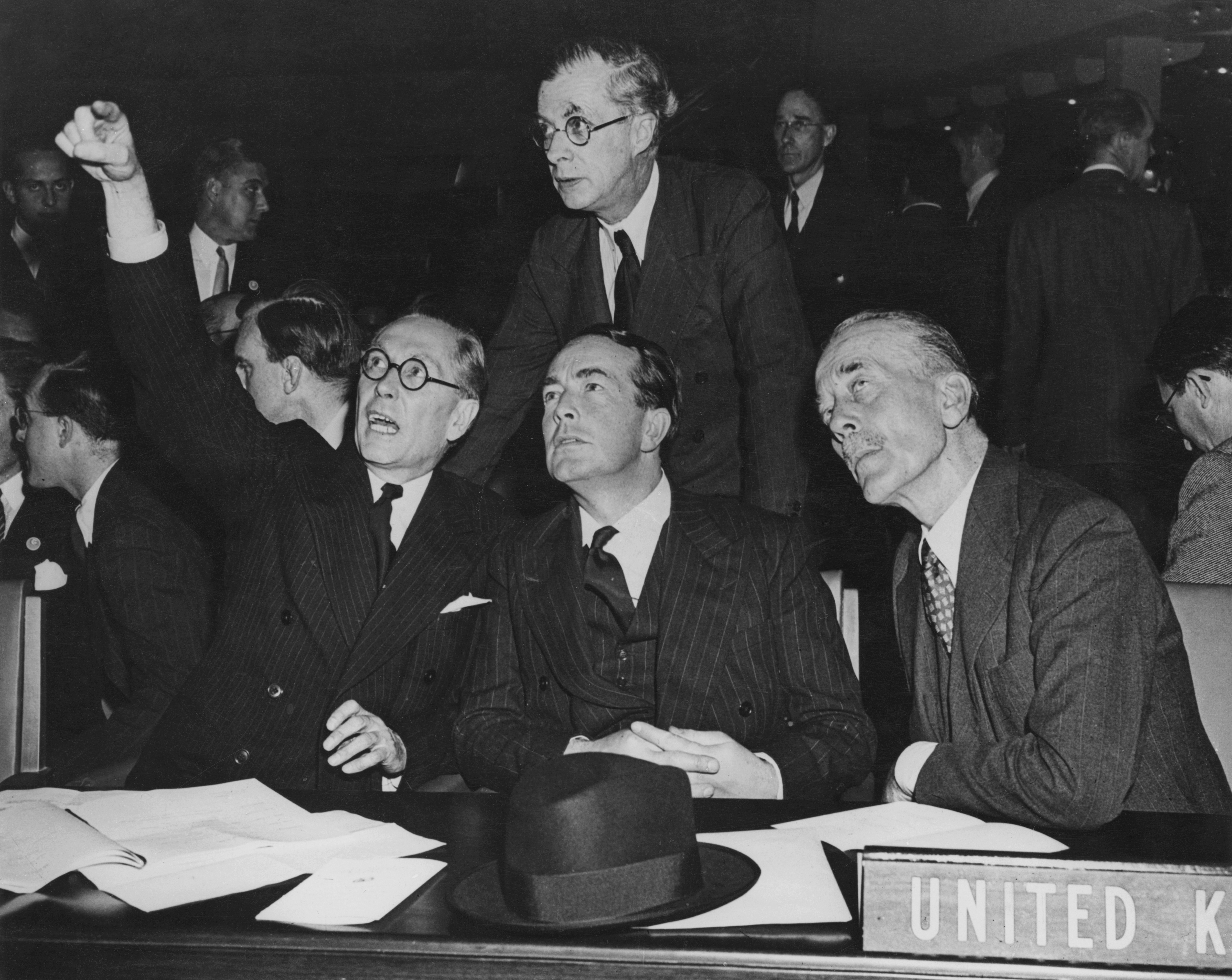 A historic black-and-white photograph from October 1946 showing Sir Philip Noel-Baker and other delegates at the United Nations General Assembly in Flushing Meadow, New York City. Four men in formal dark suits are seated at a long table labeled with a "UNITED KINGDOM" sign. One man on the left is pointing upward with his right arm while looking toward the ceiling with an open mouth, as the other three men, including one standing behind them, follow his gaze with expressions of intense focus and curiosity. A black fedora sits on the table among various scattered documents.