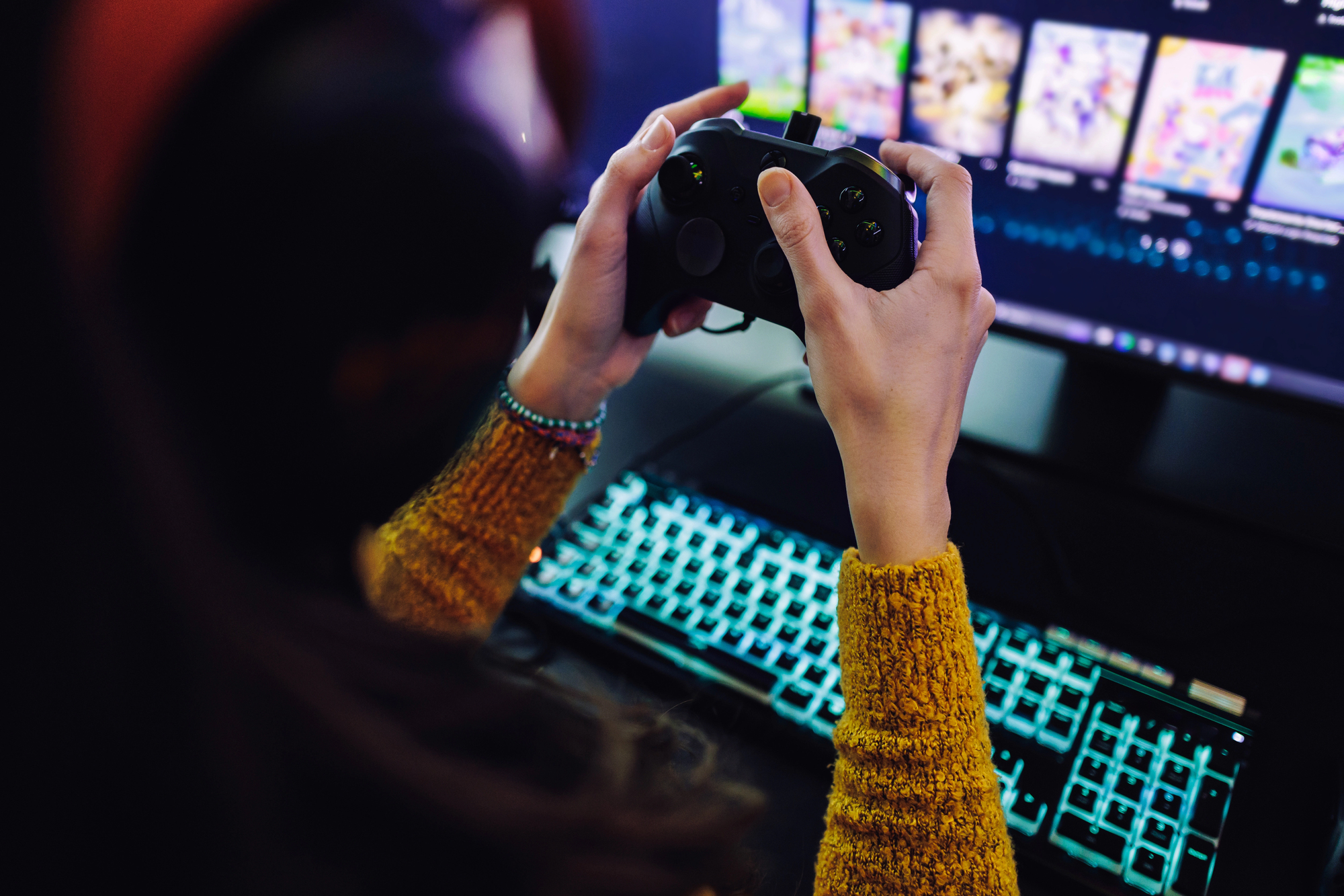 An over-the-shoulder, close-up photograph of a person playing a video game. The focus is on their hands holding a black controller, positioned above a glowing mechanical keyboard with teal backlighting. In the background, a blurred monitor displays a game selection menu or gallery.