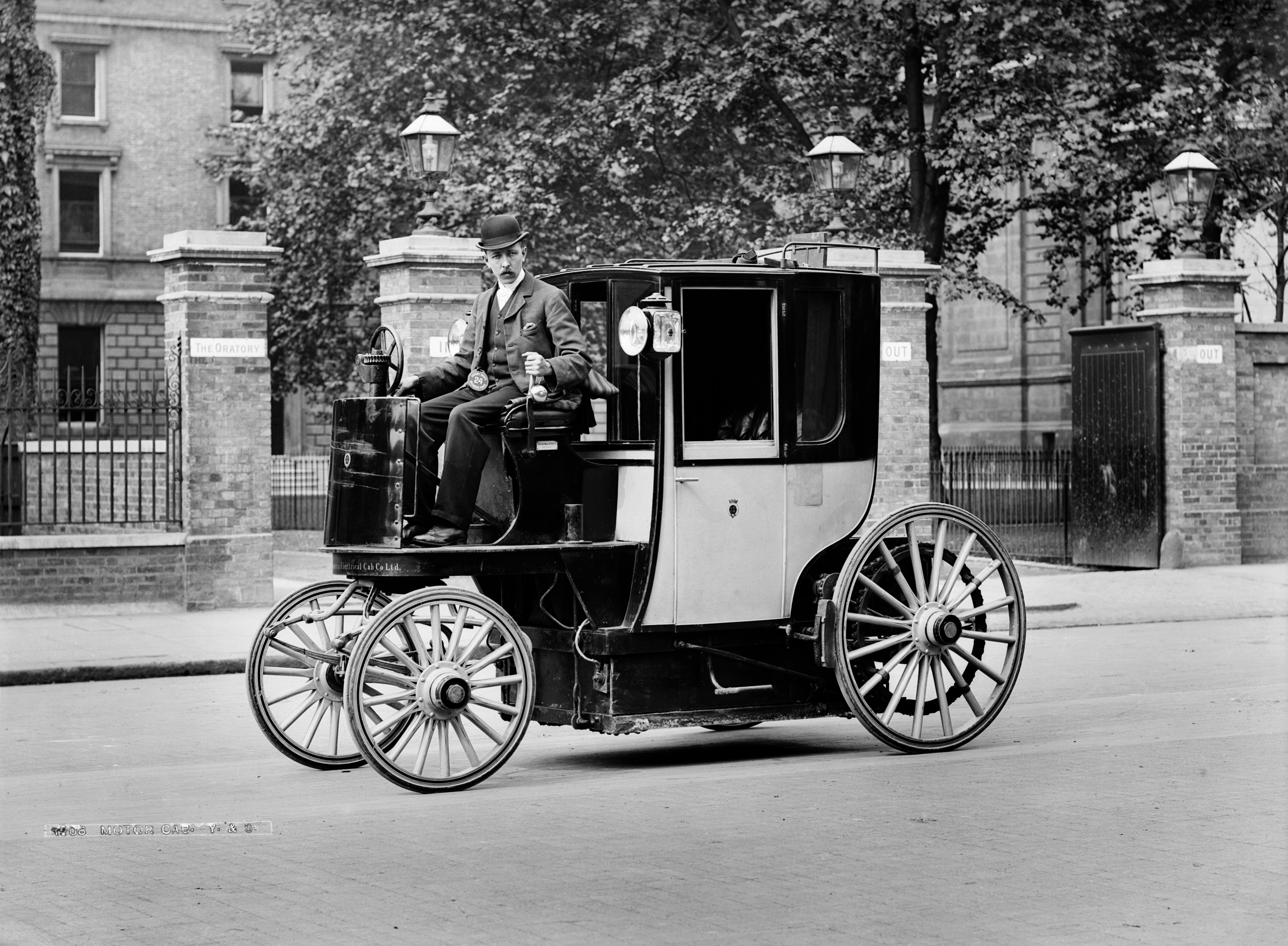 A black and white historical photograph of a Bersey Electric Cab, a vintage electric taxi, parked on a London street in front of the Brompton Oratory. The vehicle features a unique design with a large, enclosed passenger carriage at the rear and an open, elevated driver’s seat at the front. A man wearing a three-piece suit and a bowler hat sits at the steering wheel, which is a vertical column. The cab has four large, light-colored wooden-spoke wheels and two lanterns mounted near the passenger door. In the background, brick pillars with "IN" and "OUT" signs flank a gated entrance, and a large stone building is visible behind a row of trees.