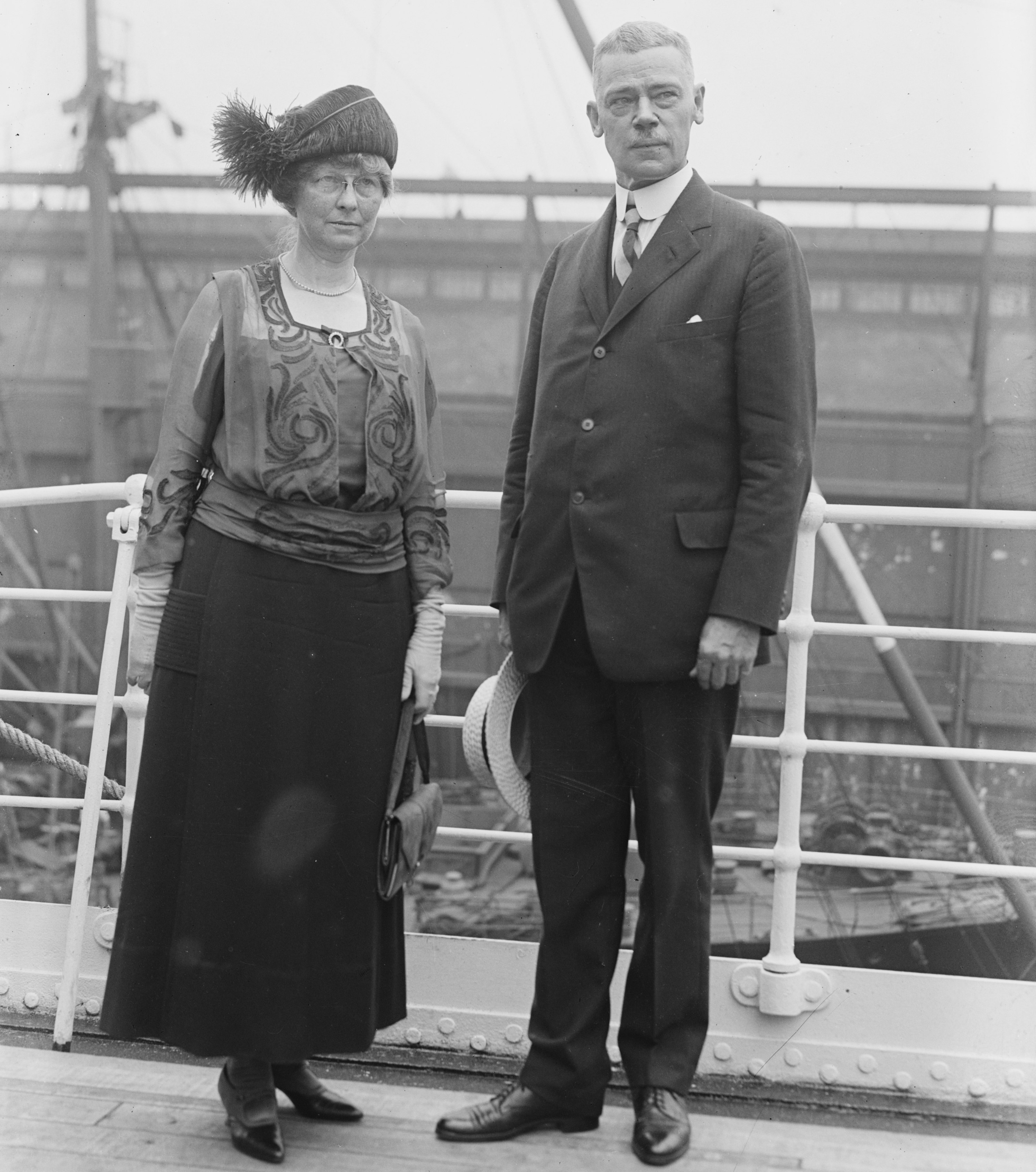 A black-and-white full-length photograph from the early 20th century featuring a man and a woman standing on the deck of a ship. The man, identified as Clinton Edgar Woods, stands on the right. He has short, light-colored hair and a mustache, wearing a dark three-piece suit with a high-collared white shirt and tie. He holds a light-colored boater hat in his right hand. The woman, his wife, stands on the left wearing a dark, floor-length skirt and a decorative, embroidered blouse with sheer sleeves. She wears a dark, wide-brimmed hat adorned with a large feather plume, spectacles, and light-colored gloves, holding a small handbag. They are positioned against a white ship railing, with the blurred industrial structures of a harbor and rigging visible in the background.