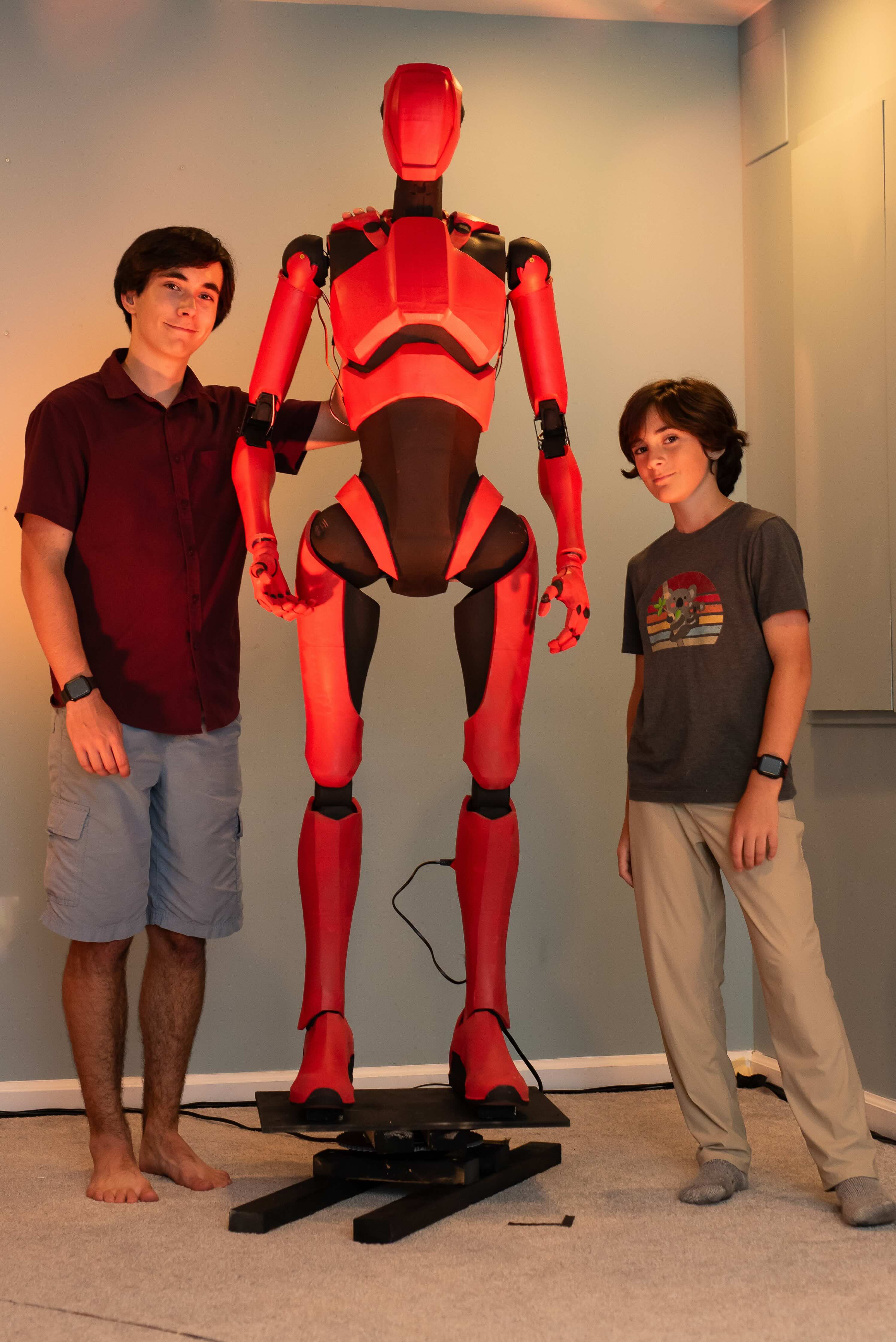 Two young men stand on either side of a tall, red and black humanoid robot prop. The robot is sleek and futuristic, mounted on a black platform in the center of a room with light-colored walls. The taller young man on the left wears a maroon button-down shirt and blue shorts, while the younger boy on the right wears a grey graphic t-shirt and tan pants.