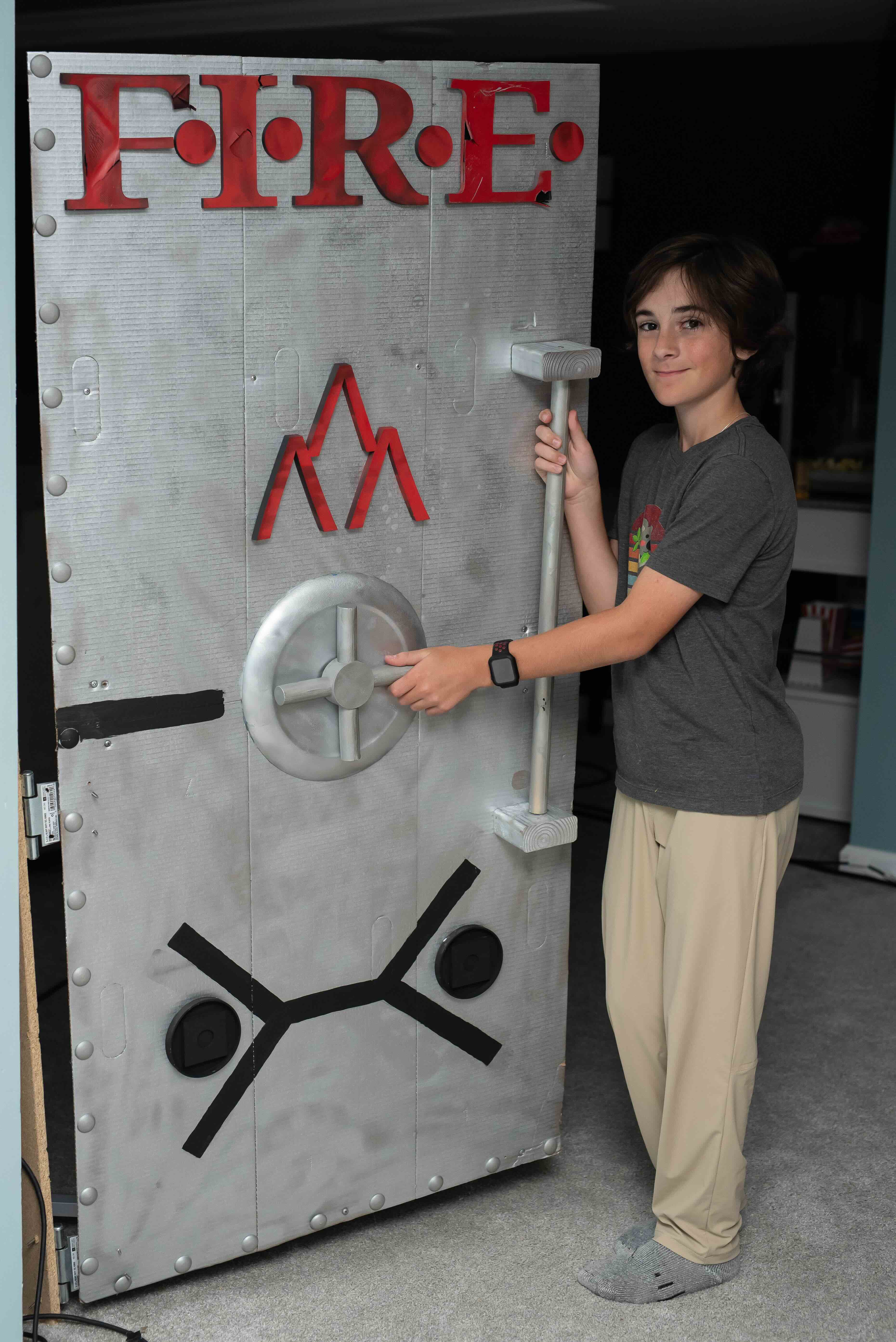 A medium shot of a smiling teenage boy with brown hair, standing next to a large, custom-built prop door designed to look like a metallic vault or industrial hatch. The door is painted silver with a weathered texture and features several distinct elements: The word "FIRE" in large, bold red letters at the top. A red mountain-like logo in the center. A silver circular wheel handle and a vertical grab bar. A black symbol at the bottom resembling a stylized person or a schematic. Silver decorative rivets lining the left and bottom edges. The boy is wearing a grey t-shirt, tan pants, and grey socks, with one hand on the wheel and the other on the grab bar. The background appears to be an indoor room with light blue walls.