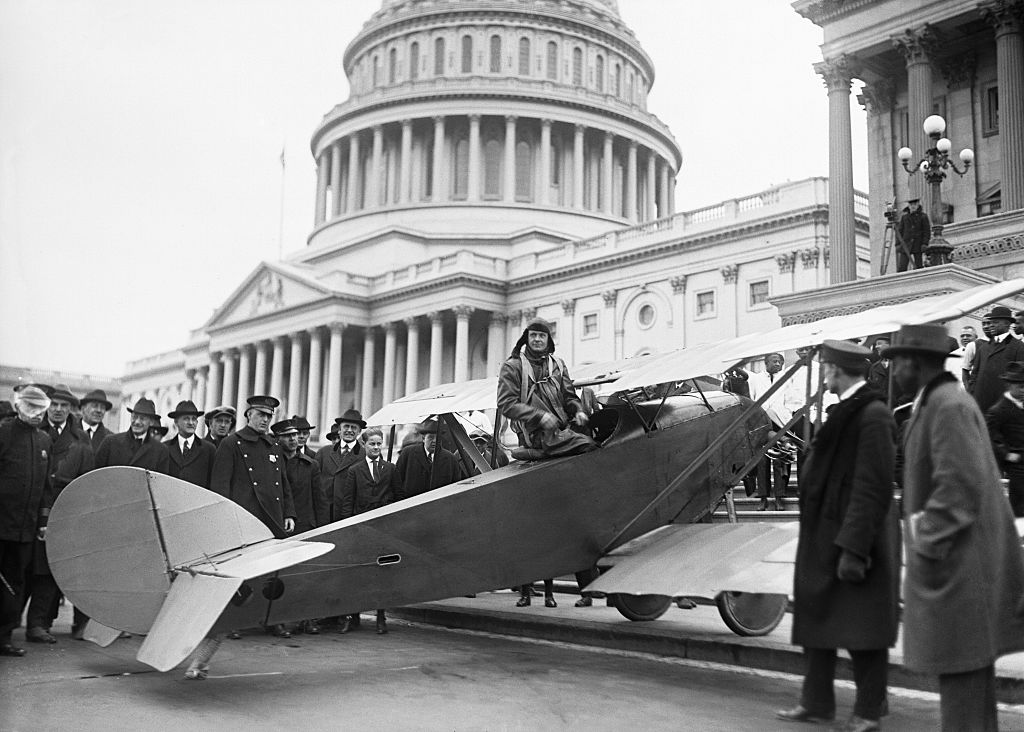 A black-and-white historical photograph of aviator Lawrence Sperry seated in the cockpit of a small biplane. He is wearing a flight cap and goggles as he sits in the stationary aircraft, which has landed directly in front of the United States Capitol building. A large crowd of men in dark overcoats and hats surrounds the plane, observing the scene against the backdrop of the Capitol's large dome and classical columns.