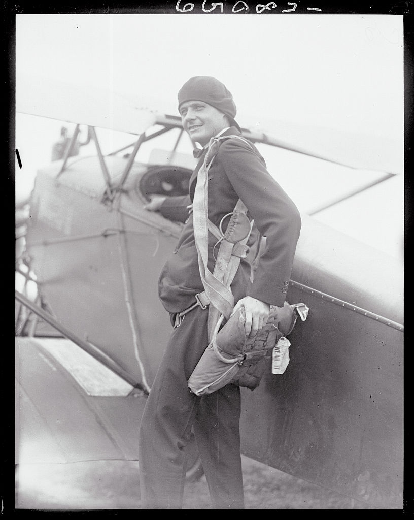 A black-and-white historical photograph shows aviator and inventor Lawrence Sperry standing beside a biplane. He is dressed in a dark suit and a soft flight cap, with a parachute pack strapped to his back and side. He is smiling and looking off-camera, with the fuselage and wing of the aircraft visible directly behind him.