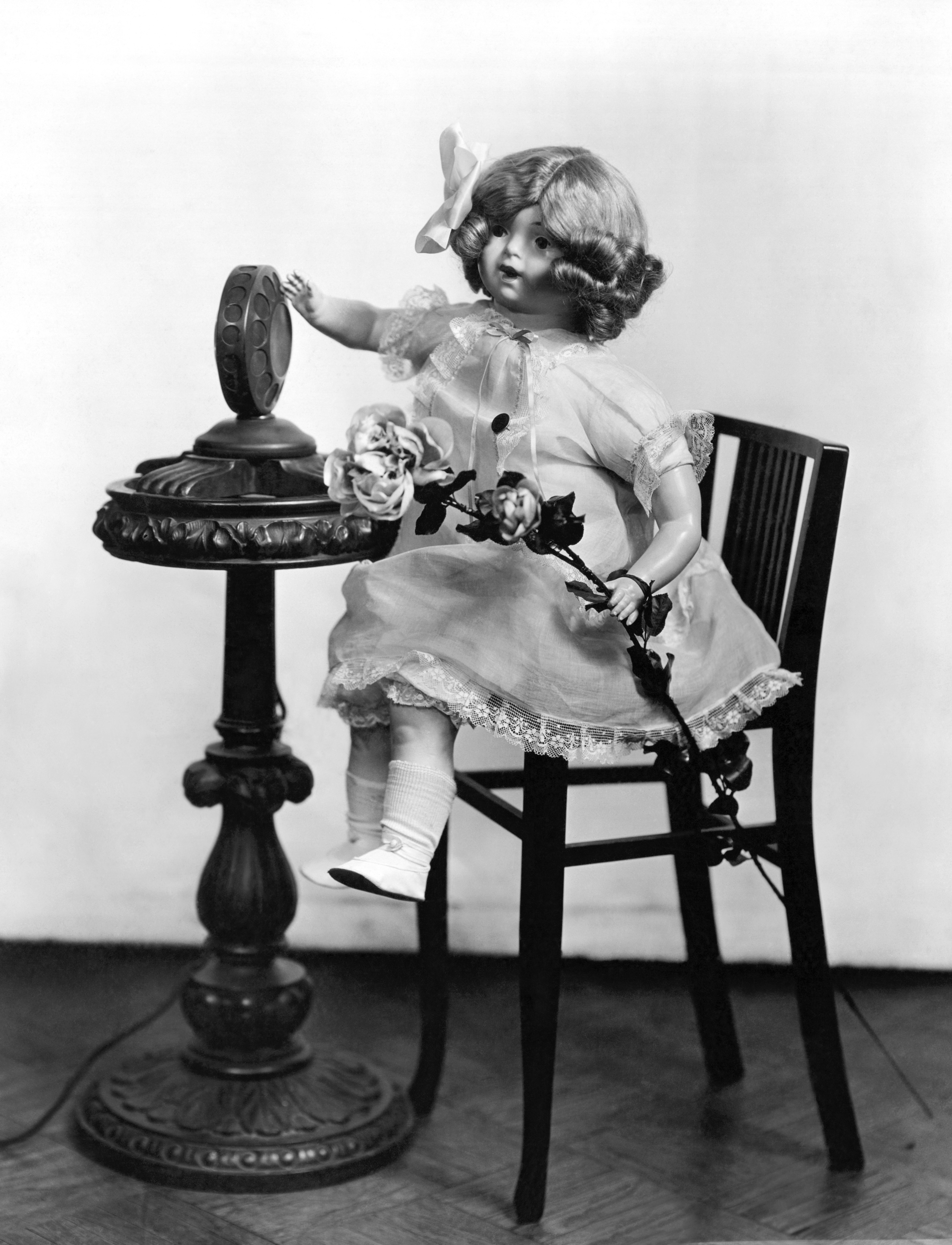 A black-and-white historical photograph of the "Radio Rose" doll, invented by Beulah Louise Henry. The doll, which has curly hair and a bow, is wearing a lace-trimmed dress and sitting on a dark wooden chair. One hand reaches out toward a circular vintage radio speaker on a pedestal table, while the other holds a long-stemmed rose.