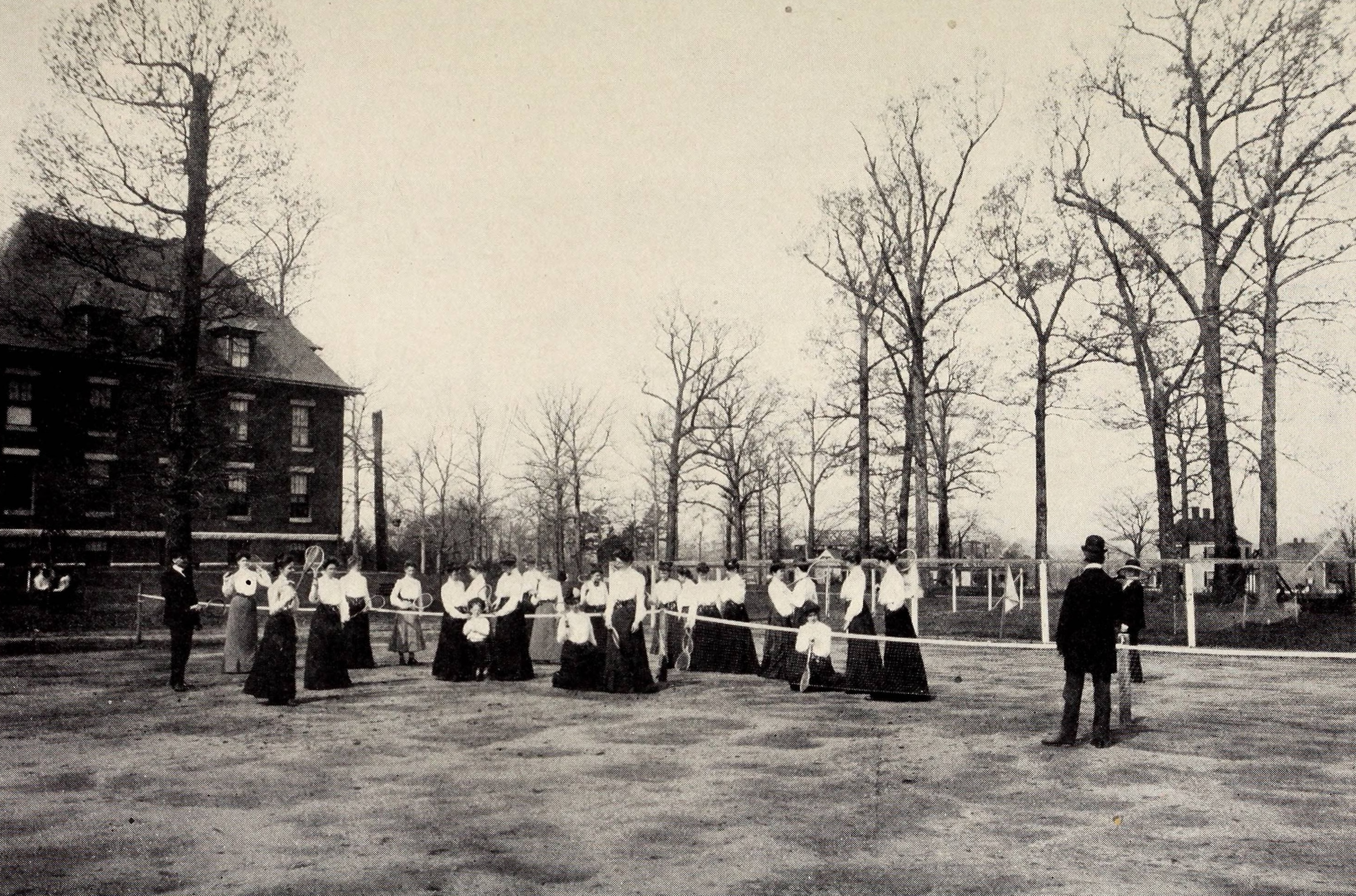 A sepia-toned historical photograph showing a large group of female students at Elizabeth College in Charlotte, North Carolina, gathered on an outdoor tennis court. The women are dressed in period-appropriate long dark skirts and white high-collared blouses, holding wooden tennis rackets. A man in a dark suit and hat stands in the foreground, looking toward the group, with a large brick college building and bare trees in the background.