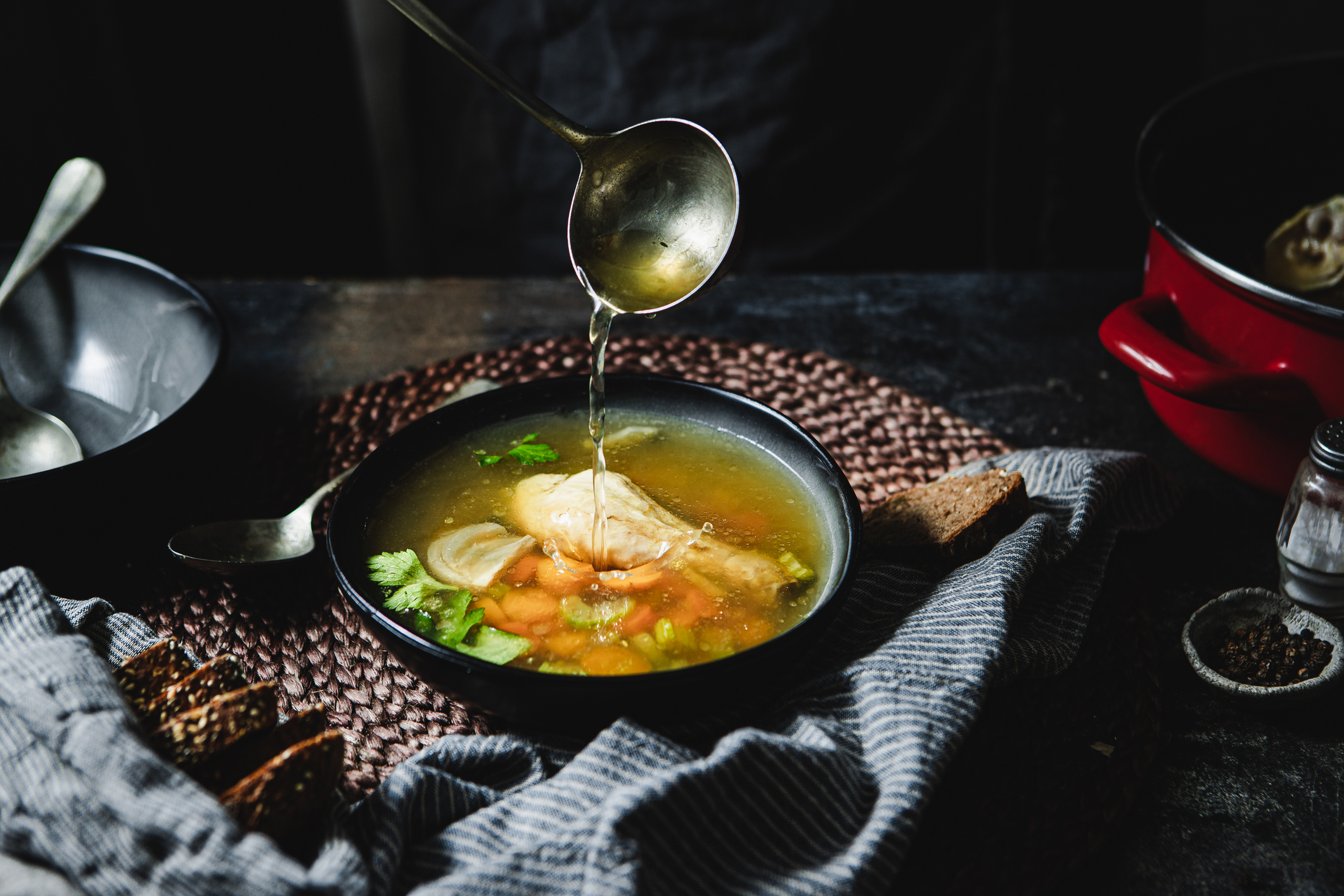 A moody, top-down photograph of a bowl of chicken soup being served. A metal ladle is captured mid-air, pouring clear, golden broth into a black bowl containing a whole chicken drumstick, sliced carrots, and celery. The bowl sits on a rustic brown woven placemat, accompanied by several slices of dark, grainy bread on a blue and white striped linen napkin. The scene is set against a dark, textured background, with a red cooking pot visible in the upper right.