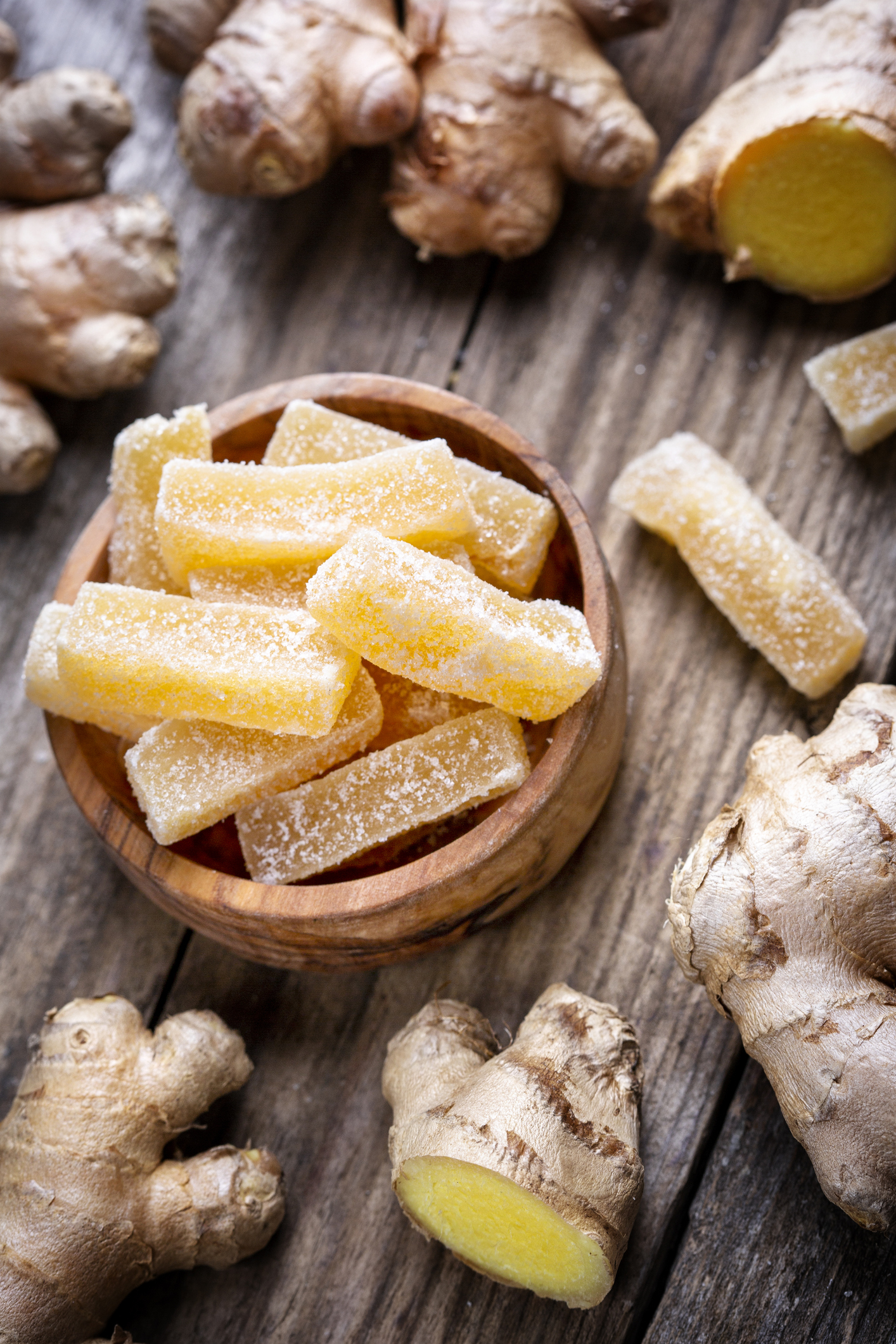 A top-down, close-up photograph of several whole, raw ginger roots scattered across a rustic wooden surface. In the center, a small round wooden bowl is filled with rectangular, sugar-coated ginger chews. One of the raw ginger roots in the foreground is sliced open to reveal its bright yellow interior.