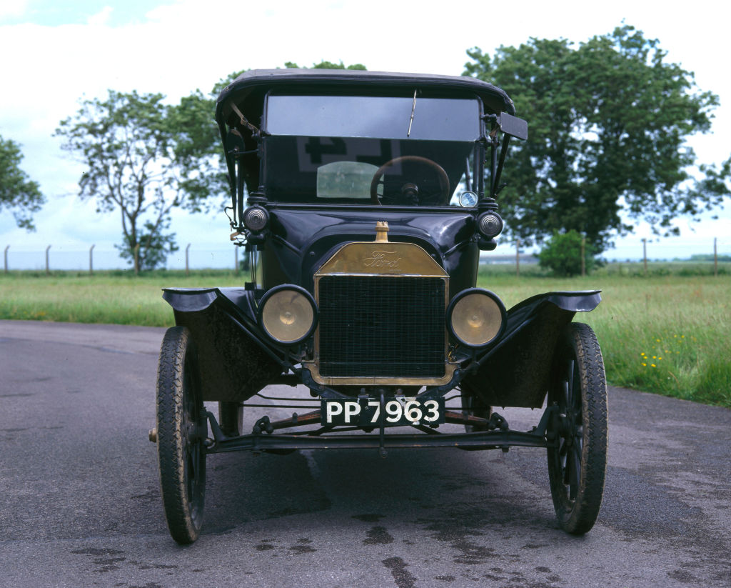A head-on photograph of a vintage black 1916 Ford Model T four-seat tourer motor car. The vehicle features large circular headlights, a brass-framed radiator with the Ford logo, and a British license plate reading PP 7963. It is parked on a paved road in front of a green field under a pale sky.