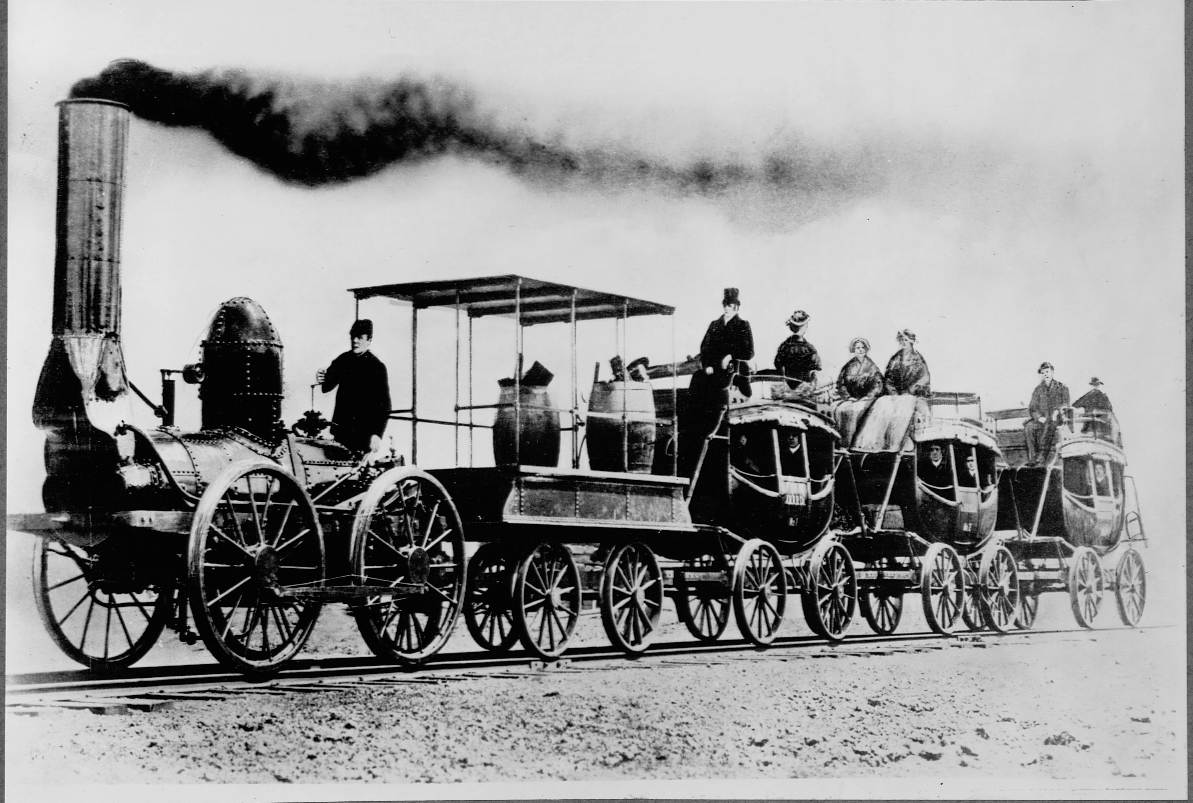 Black and white image showing an open-air DeWitt Clinton's locomotive pulls stagecoaches converted for use as passenger cars. Several passengers ride in and on top of the stagecoaches.