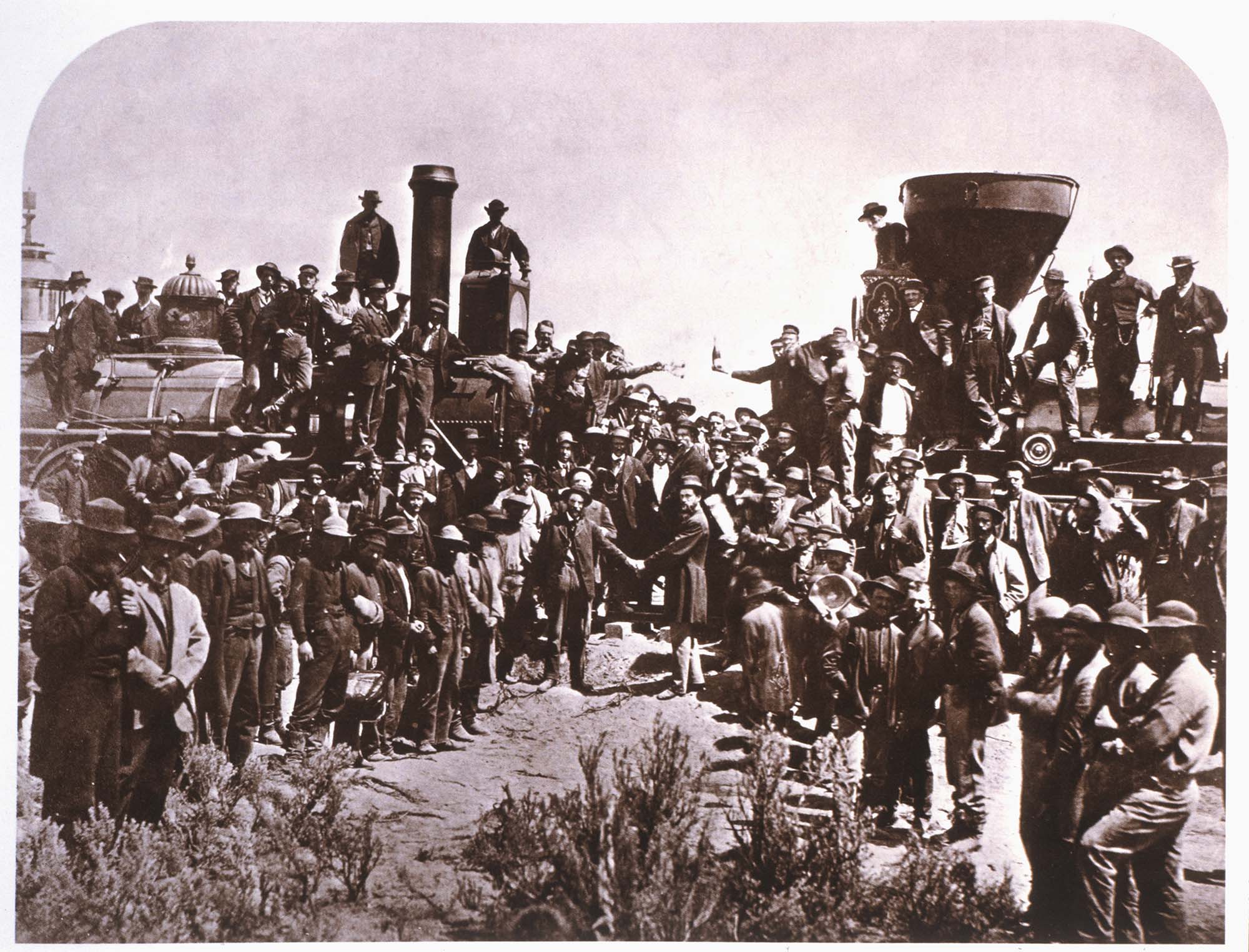 Sepia-colored image of crowd at the completion of the Trans-Continental Railroad in Pomontory, Utah, 1869. Many men celebrate and two men in the center shake hands.