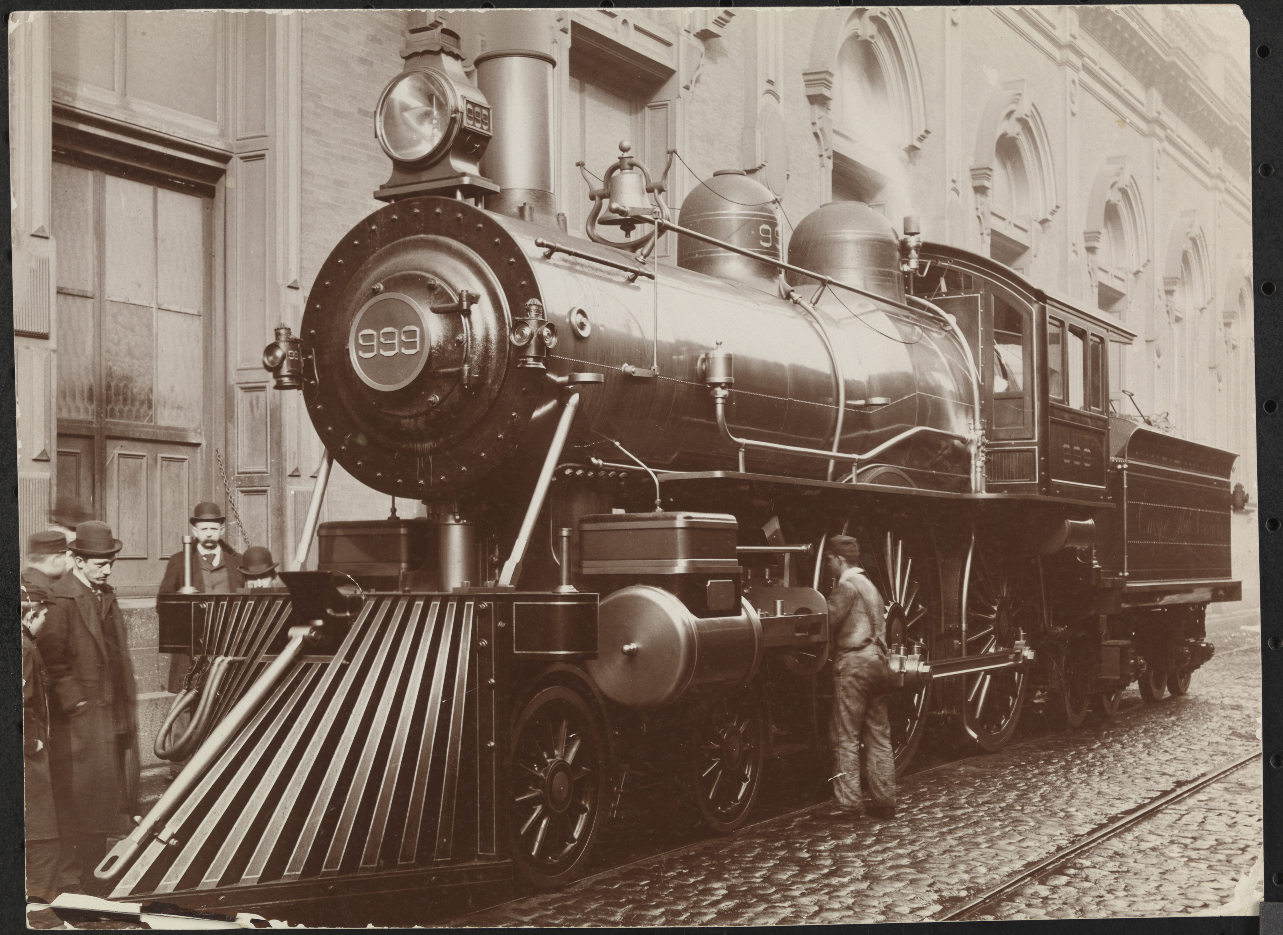 Black and white photo. Railroad locomotive--#999--on tracks at what may be the New York Central Railroad building with men in foreground, New York, New York, late 1890s. 