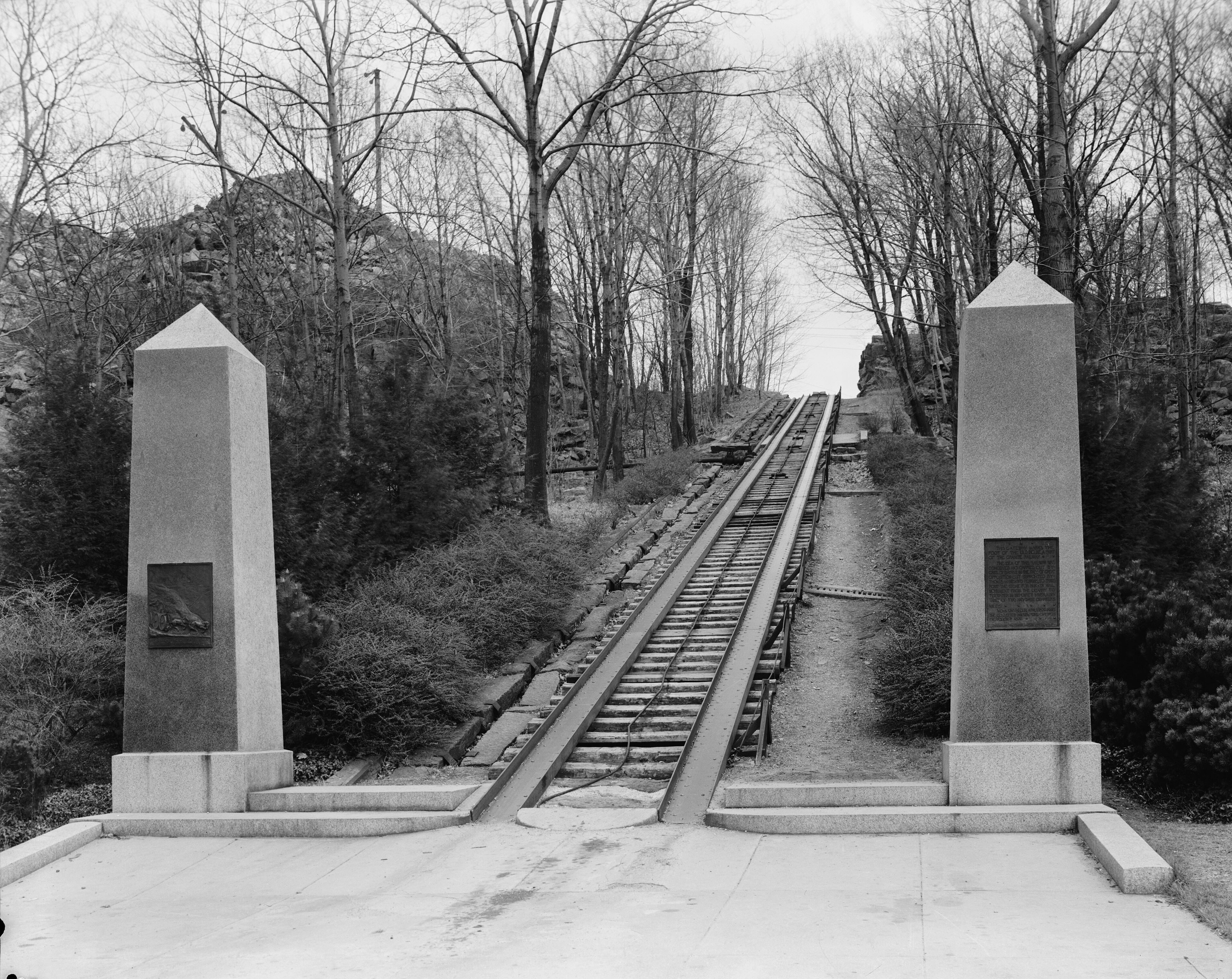 A black-and-white historical photograph shows the preserved site of the Granite Railway in Quincy, Massachusetts, one of the first railroads in the United States. Two large, identical stone obelisks with commemorative bronze plaques frame the entrance to a steep incline plane. The railway tracks, consisting of iron-capped granite rails, extend upward into a wooded area flanked by bare trees. The foreground shows a flat stone platform leading to the base of the track.