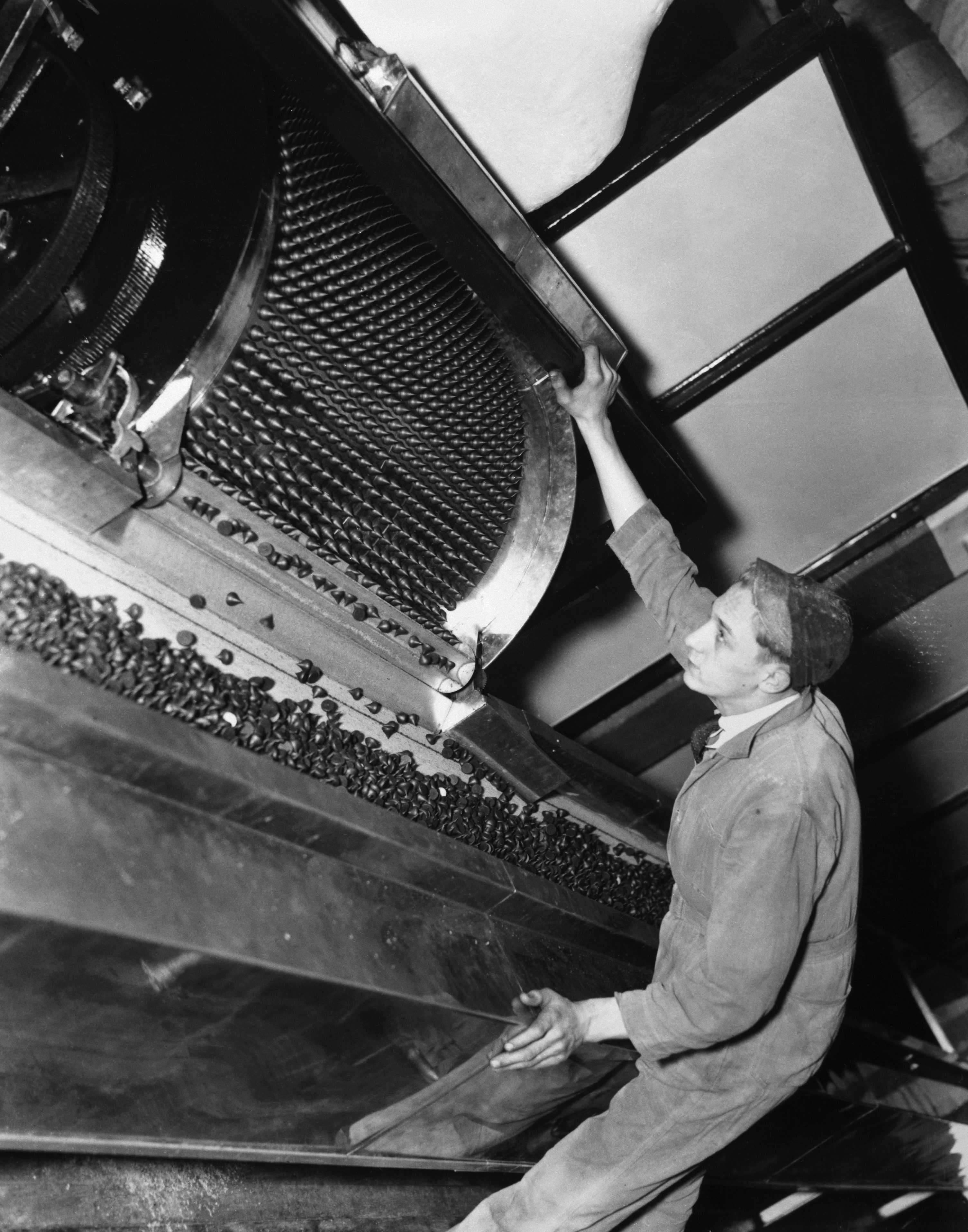 A black-and-white historical photograph captures a factory worker monitoring a large industrial machine producing Hershey's Kisses. The worker, dressed in light-colored overalls and a cap, leans forward to inspect a rotating drum filled with hundreds of the iconic teardrop-shaped chocolates. Below the drum, a conveyor belt is piled high with finished Kisses moving through the production line.