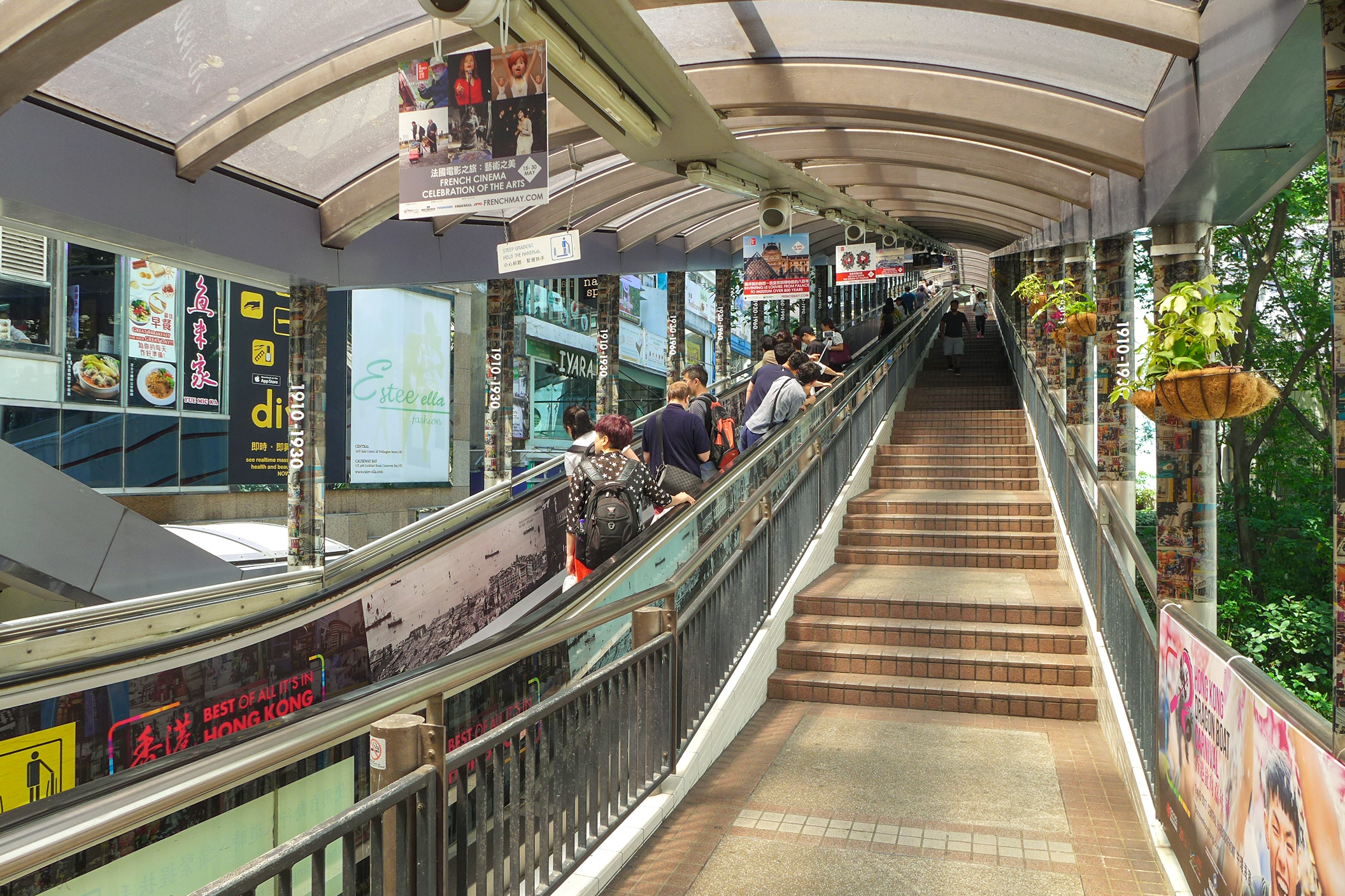 The interior of the Central-Mid-Levels Escalator in Hong Kong. A moving sidewalk goes up the left side of a covered elevated structure with stairs to the right. There are ads along the escalator and trees can be seen outside.