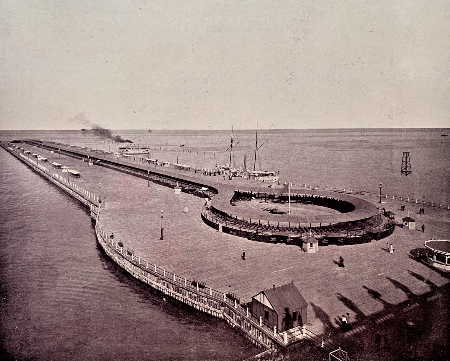 A high-angle, sepia-toned historical photograph shows the moving sidewalk at the 1893 World's Columbian Exposition in Chicago. The wooden pier extends far into Lake Michigan, featuring a long, looped mechanical walkway with rows of benches for passengers. Several steamships and sailboats are docked alongside the pier, and a few fairgoers are visible walking across the expansive wooden deck.