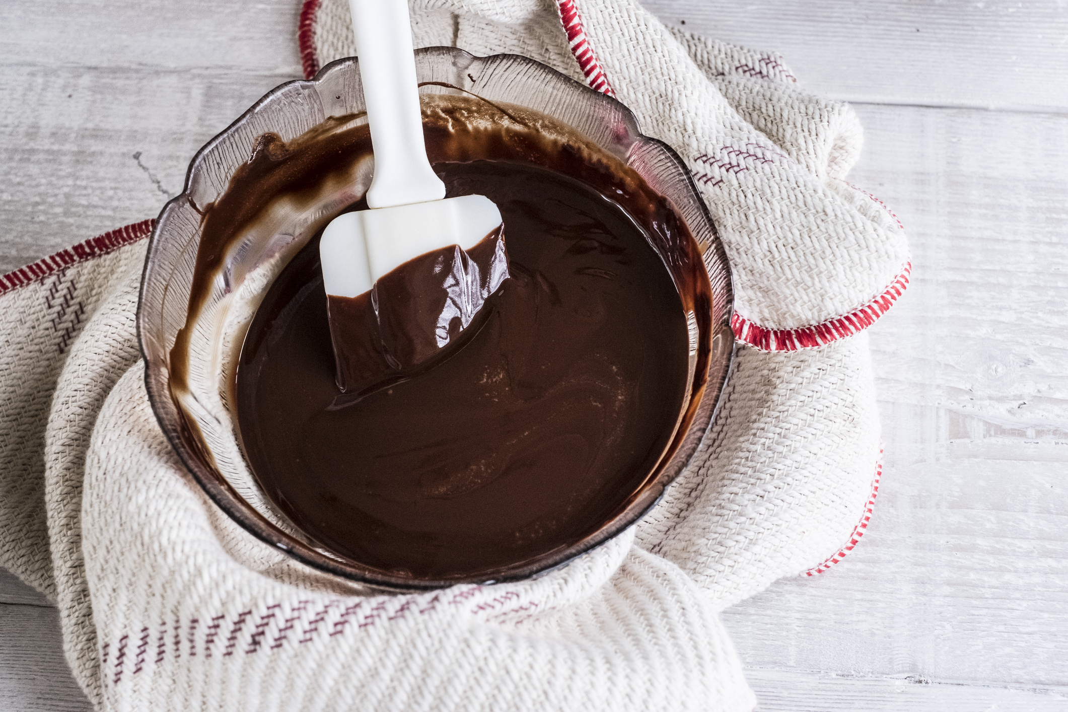 A glass, petal bowl of chocolate brownie dough being stirred by a white spatula. The bowl rests on a white towel on a white-washed wooden surface.