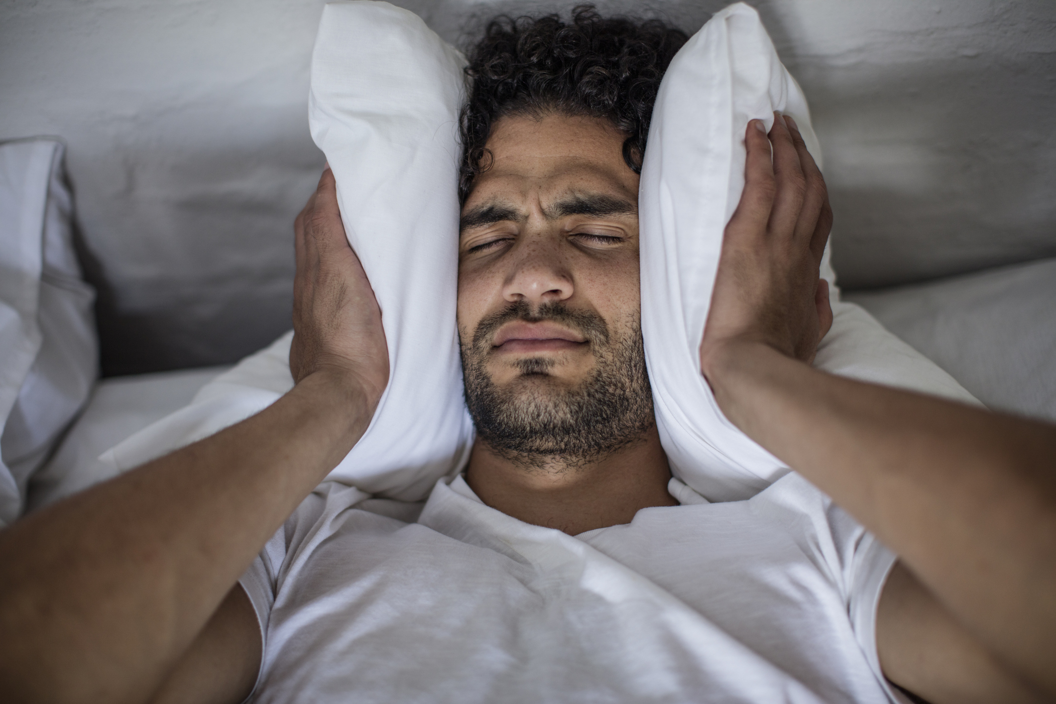 Young mixed race man presses his white pillow around his ears presumably from a loud noise disrupting his sleep.