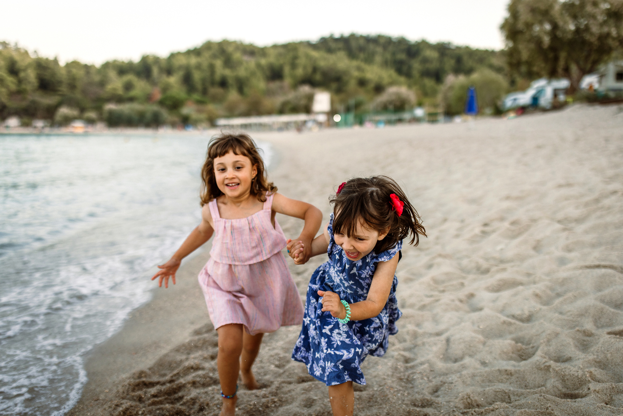 A medium-shot, candid photograph shows two young, barefoot girls holding hands and running along a sandy beach at sunset. One girl wears a light pink sleeveless dress, while the other is in a blue floral dress with red bows in her hair. They are both laughing and smiling as they run near the water's edge, with a calm sea on the left and a lush, tree-covered hill in the background.