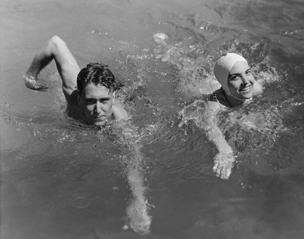 Black and white vintage 1940s white couple swimming in swimming pool. The woman wears a swimming cap.